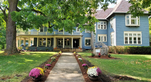 A large blue Victorian-style house with a porch, surrounded by green trees and colorful flower beds along the pathway.