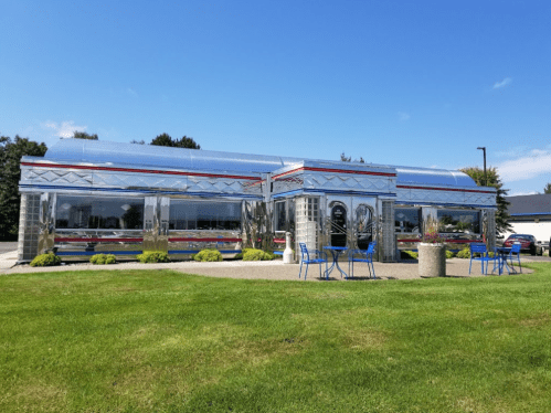 A shiny, retro-style diner with chrome exterior and colorful chairs on a grassy area under a clear blue sky.