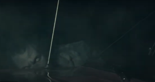 A shadowy figure with glowing eyes is partially obscured by fog, peering from behind a car hood.