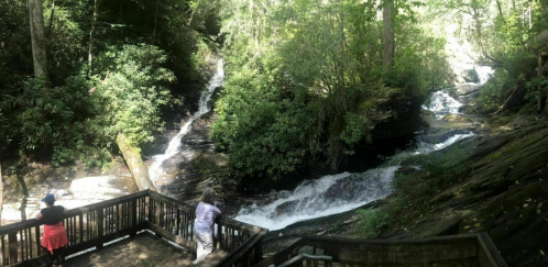Two people stand on a wooden deck overlooking a serene waterfall surrounded by lush greenery.