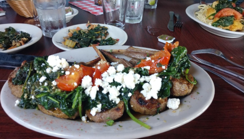 A plate of grilled meat topped with spinach, tomatoes, and feta cheese, surrounded by side dishes and drinks.