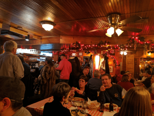 A bustling restaurant with wooden decor, red lights, and patrons enjoying meals at checkered tables.