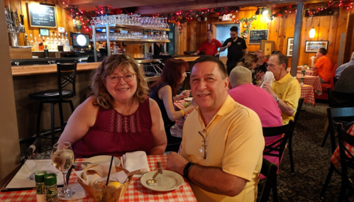 A couple smiles at the camera while dining in a cozy restaurant decorated with lights and festive decor.