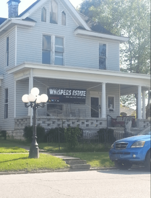 A large, light blue house with a "Whispers Estate" banner and a vintage streetlamp in front.