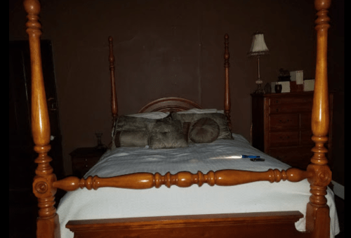 A wooden four-poster bed with white bedding and decorative pillows in a dimly lit room. A lamp and dresser are visible.