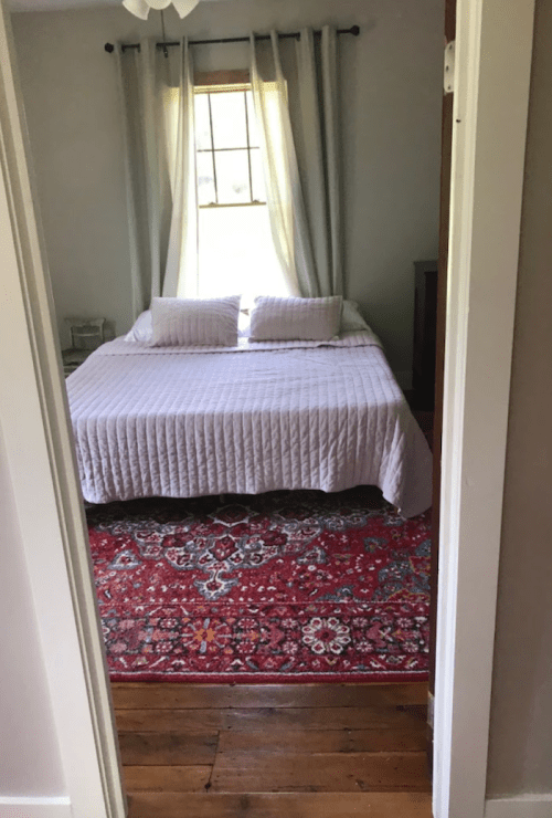 A cozy bedroom with a bed covered in a light quilt, two pillows, and a colorful rug, seen through an open doorway.
