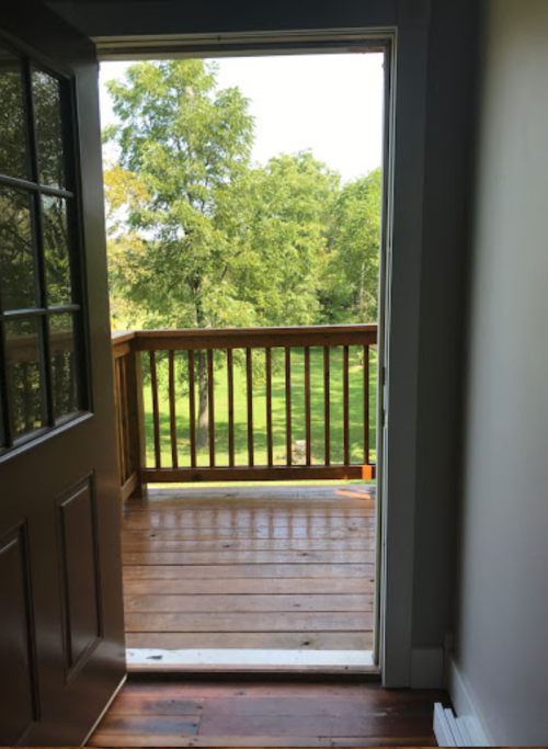 A doorway opens to a wooden balcony overlooking a green landscape with trees.