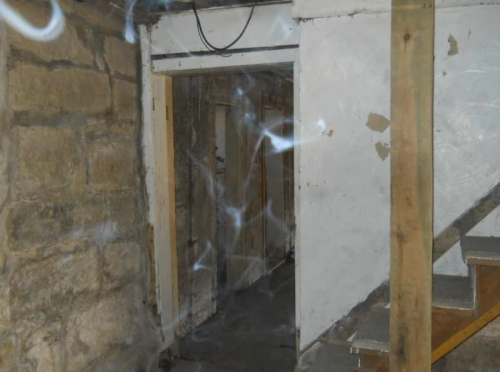 A dimly lit basement with stone walls, a wooden staircase, and an open doorway leading to another room.