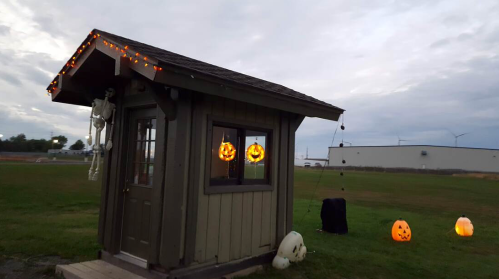 A small cabin decorated with Halloween lights and pumpkins, set against a cloudy sky and grassy field.
