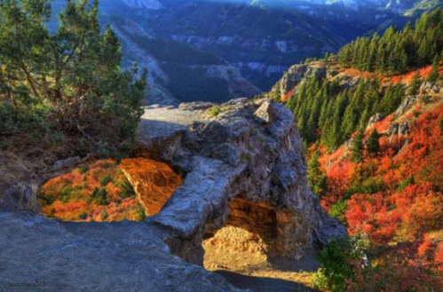 A rocky archway surrounded by vibrant autumn foliage and mountains in the background, illuminated by soft sunlight.