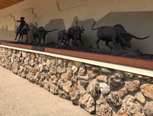 A bronze sculpture of a cowboy on horseback leading a herd of longhorn cattle against a stone wall backdrop.