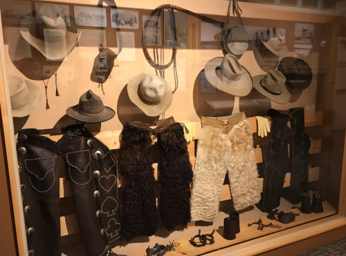 Display of cowboy hats, chaps, gloves, and gear in a museum exhibit showcasing Western attire.