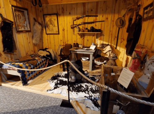 A rustic wooden room with a bed, table, and various historical artifacts, including guns and clothing.