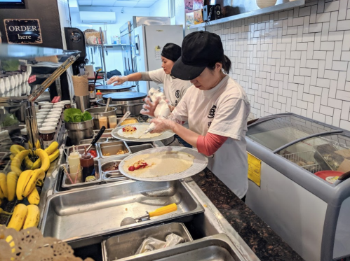 Two women prepare food in a busy kitchen, assembling crepes with various ingredients and toppings.