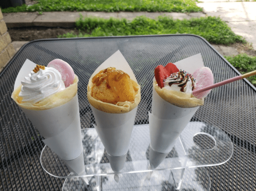 Three crepes in cones: one with whipped cream, one with fruit, and one with strawberries and sprinkles, on a table.