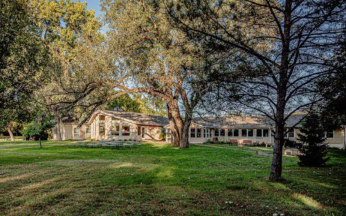A spacious building surrounded by trees and a green lawn, with a clear blue sky in the background.