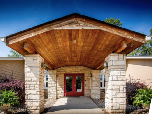 Entrance of a building featuring a wooden roof, stone pillars, and red double doors, surrounded by greenery.