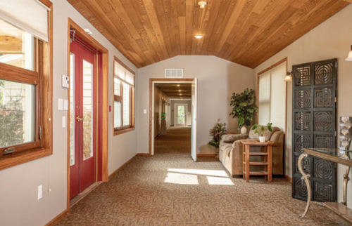 A cozy hallway with wooden ceiling, red door, and a seating area, leading to multiple doorways and greenery.