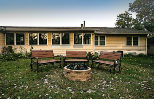 A cozy backyard with four benches around a stone fire pit, set against a house with large windows.