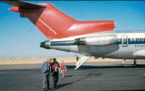 A red-tailed airplane on a runway with three people walking nearby, set against a clear blue sky.