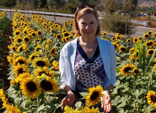 A woman stands among vibrant sunflowers, smiling and holding a sunflower, with a scenic background of trees and a road.