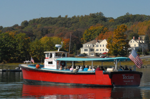 A red boat named "Teciani" with passengers, cruising on calm water near colorful autumn trees and a white house.