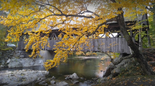 A wooden covered bridge surrounded by vibrant autumn leaves over a calm river with rocks.
