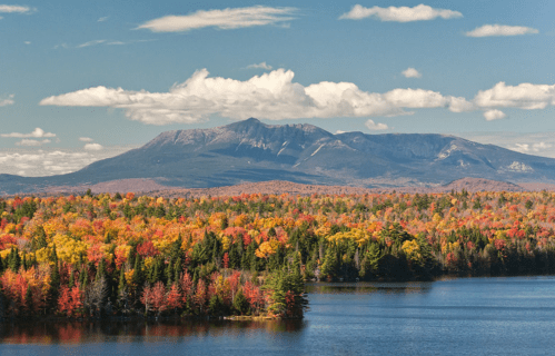 A vibrant autumn landscape featuring colorful trees, a serene lake, and a mountain under a blue sky with fluffy clouds.