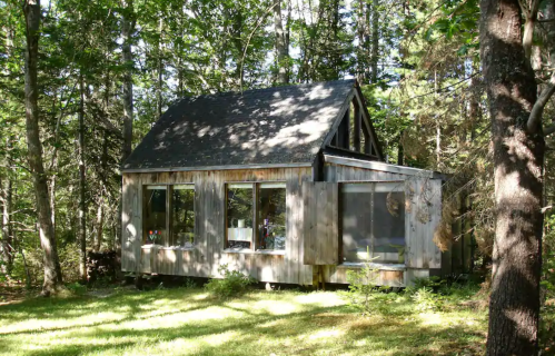 A rustic wooden cabin surrounded by trees, featuring large windows and a sloped roof in a serene forest setting.