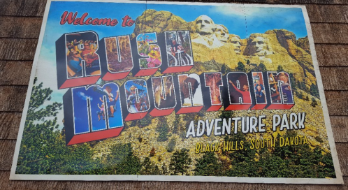 A colorful sign reading "Welcome to Rush Mountain Adventure Park" with a mountain backdrop in Black Hills, South Dakota.