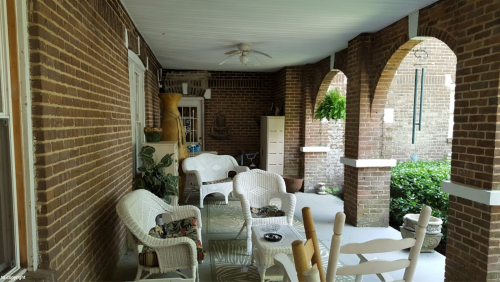 A cozy porch with wicker furniture, potted plants, and brick walls, featuring arches and a ceiling fan.