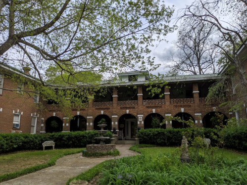 A brick building with arched entrances, surrounded by greenery and a small fountain in the courtyard.