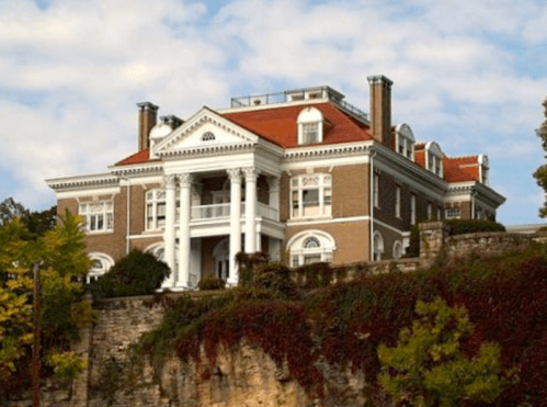 A large, elegant mansion with a red roof and white columns, set against a cloudy sky and surrounded by greenery.