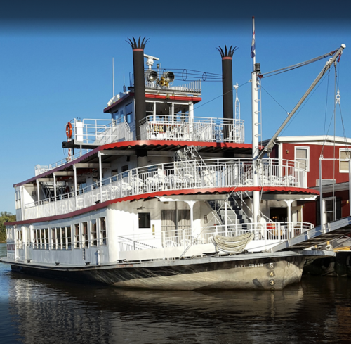 A large, white riverboat with red accents and black smokestacks docked on calm water under a clear blue sky.