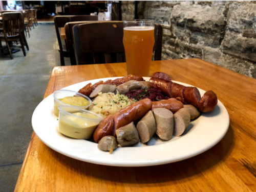 A plate of assorted sausages with sauerkraut and mustard, accompanied by a glass of beer on a wooden table.