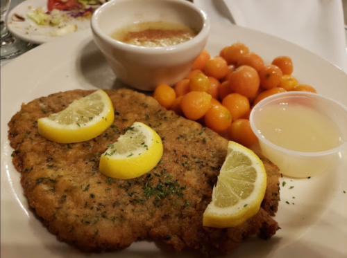 A plate featuring breaded chicken with lemon slices, a side of cantaloupe balls, and a small bowl of sauce.