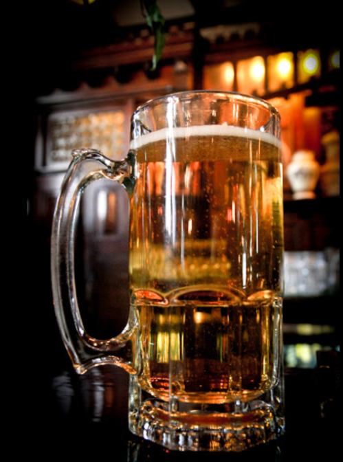 A close-up of a frothy glass of beer on a dark bar counter, with warm lighting in the background.