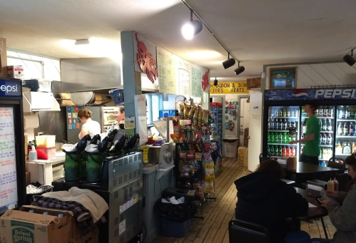 A cozy convenience store with a kitchen area, shelves of snacks, and customers seated at tables.