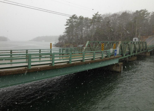 A green bridge over a river in snowy weather, surrounded by trees and a misty landscape.