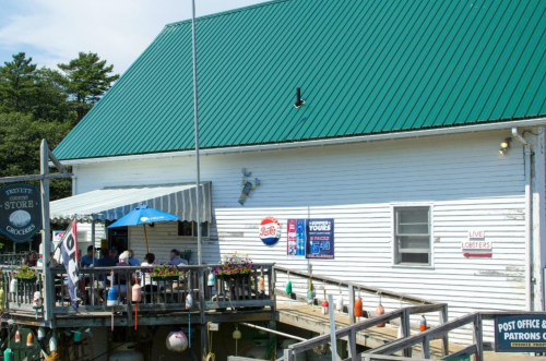 A coastal store with a green roof, featuring outdoor seating and signs for live lobsters and a post office.