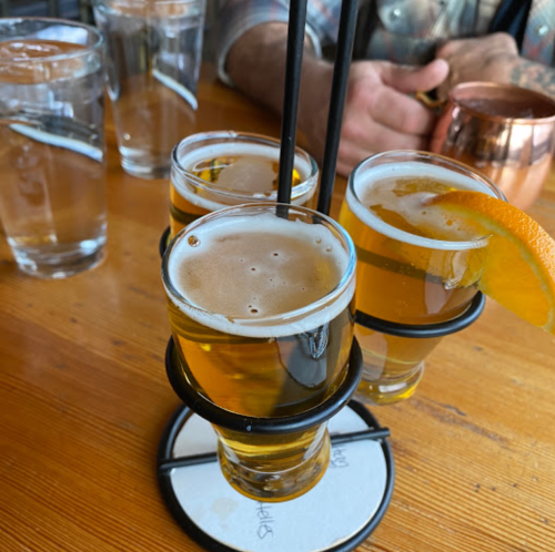 Three small glasses of beer with a slice of orange, served on a wooden table alongside water glasses.