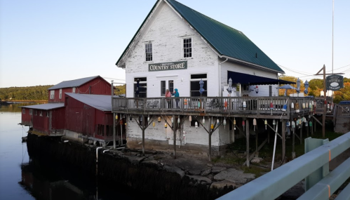 A white country store on stilts by the water, with people on the deck and red buildings in the background.