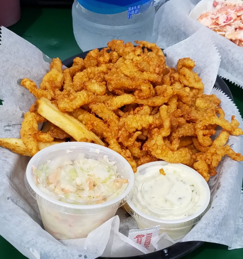 A basket of crispy fried clam strips with fries, accompanied by coleslaw and dipping sauces.