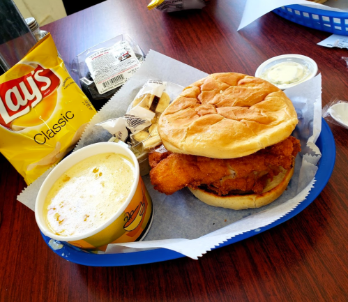 A fried chicken sandwich on a bun, served with a side of soup, chips, and a small container of sauce on a blue tray.