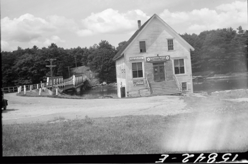 A vintage black-and-white photo of a building by a river, with a bridge and trees in the background.