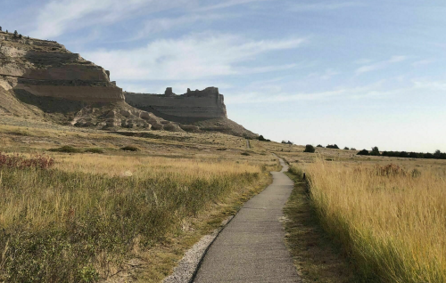 A winding path leads through tall grass towards rocky cliffs under a clear blue sky.