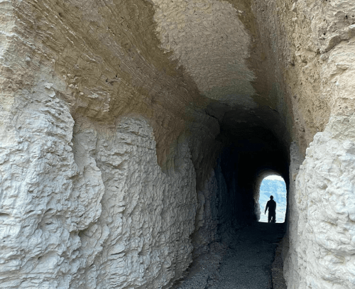 A silhouette of a person stands inside a rocky tunnel, with light visible at the far end.