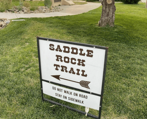 Sign for Saddle Rock Trail with instructions: "Do not walk on road, stay on sidewalk." Green grass in the background.