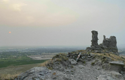 A rocky outcrop overlooks a hazy landscape at sunset, with fields and a distant town visible below.