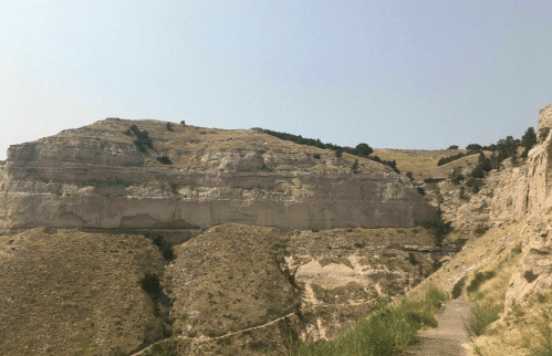 A rocky landscape with layered cliffs and sparse vegetation under a clear sky. A path winds through the terrain.